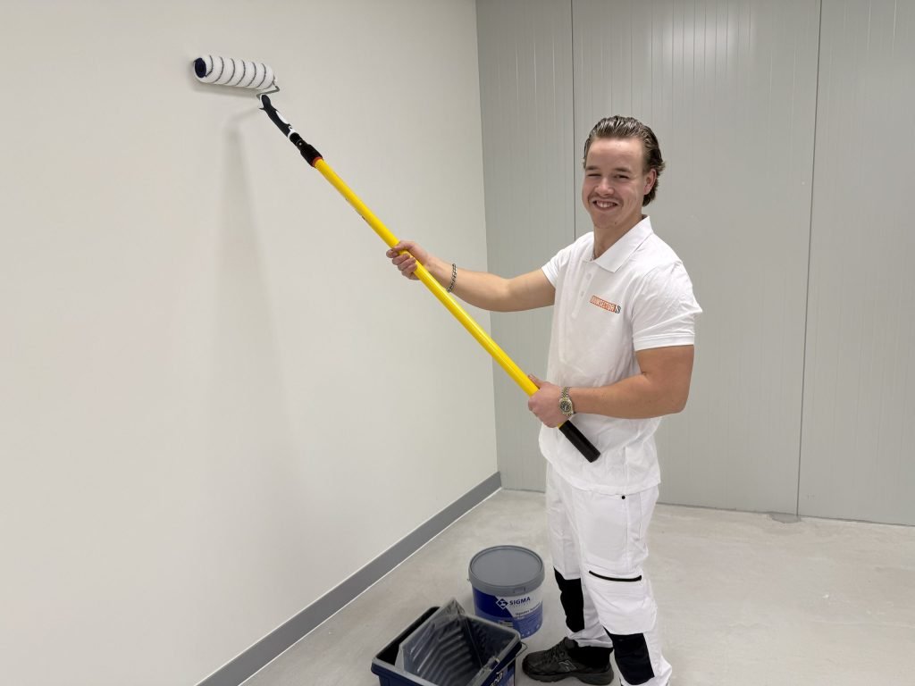 Lachende schilder in wit werkpak rolt een witte wand, met een emmer Sigma-verf en een verfbak op de vloer