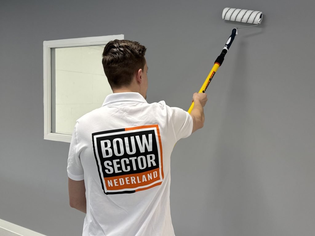 Schilder met Bouwsector-logo op de rug brengt grijze muurverf aan naast een raamkozijn