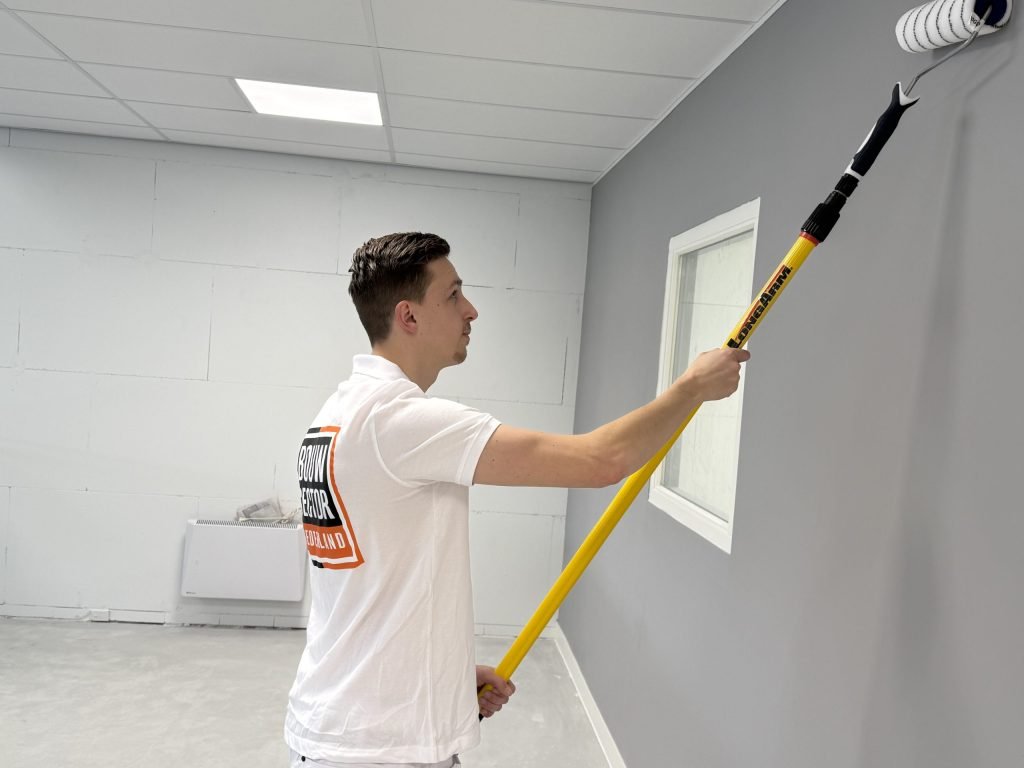 Schilder in wit poloshirt zet met een telescoopsteel een grijze muur in de verf naast een raam