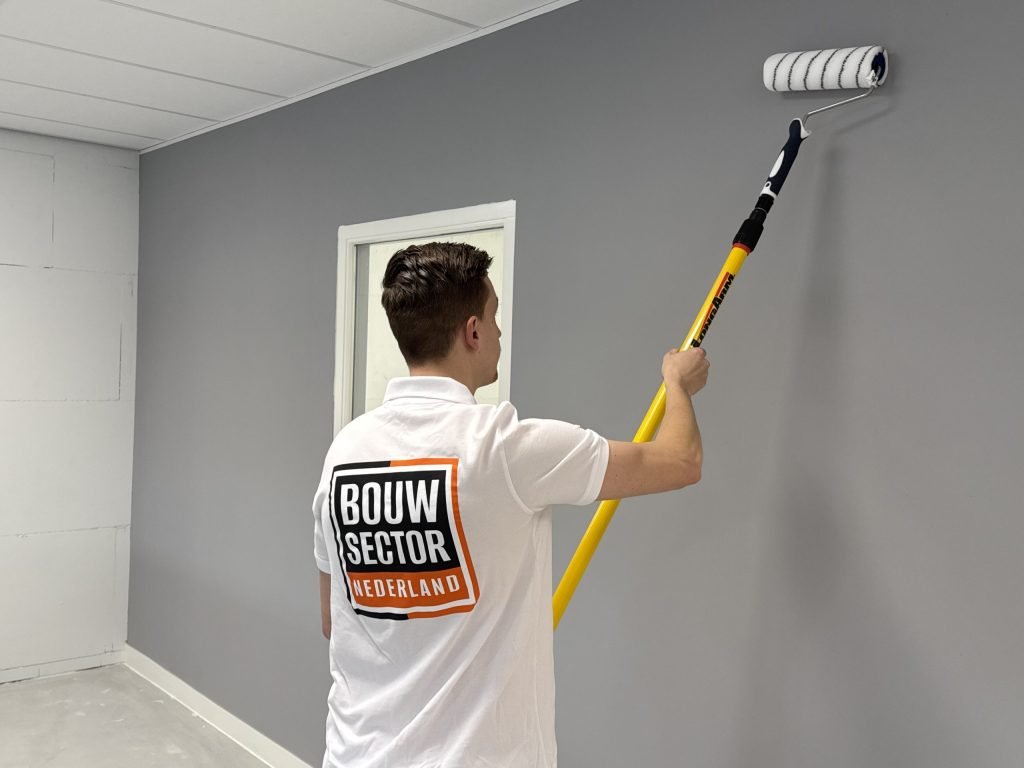Schilder rolt een grijze wand in een ruimte met een systeemplafond en witte plinten