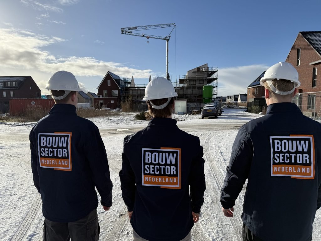 Drie medewerkers van Bouwsector Nederland in donkere jas en witte helm op een besneeuwde straat met bakstenen huizen
