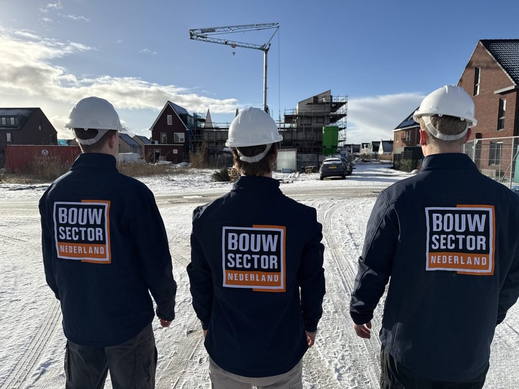 Drie mensen van Bouwsector Nederland met witte helm en bedrijfsjas staan met de rug naar de camera op een besneeuwde bouwweg bij nieuwe woningen